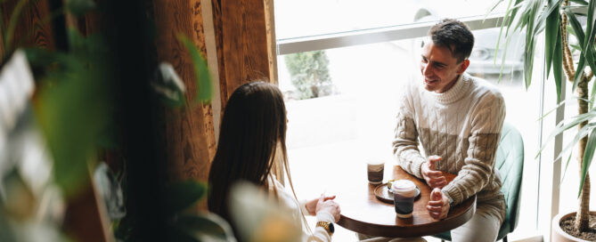 A man and woman are seated at a table engaging in conversation. They appear to be in deep discussion, leaning towards each other. The table is bare except for a few cups and plates. The man gestures with his hands while the woman listens intently.