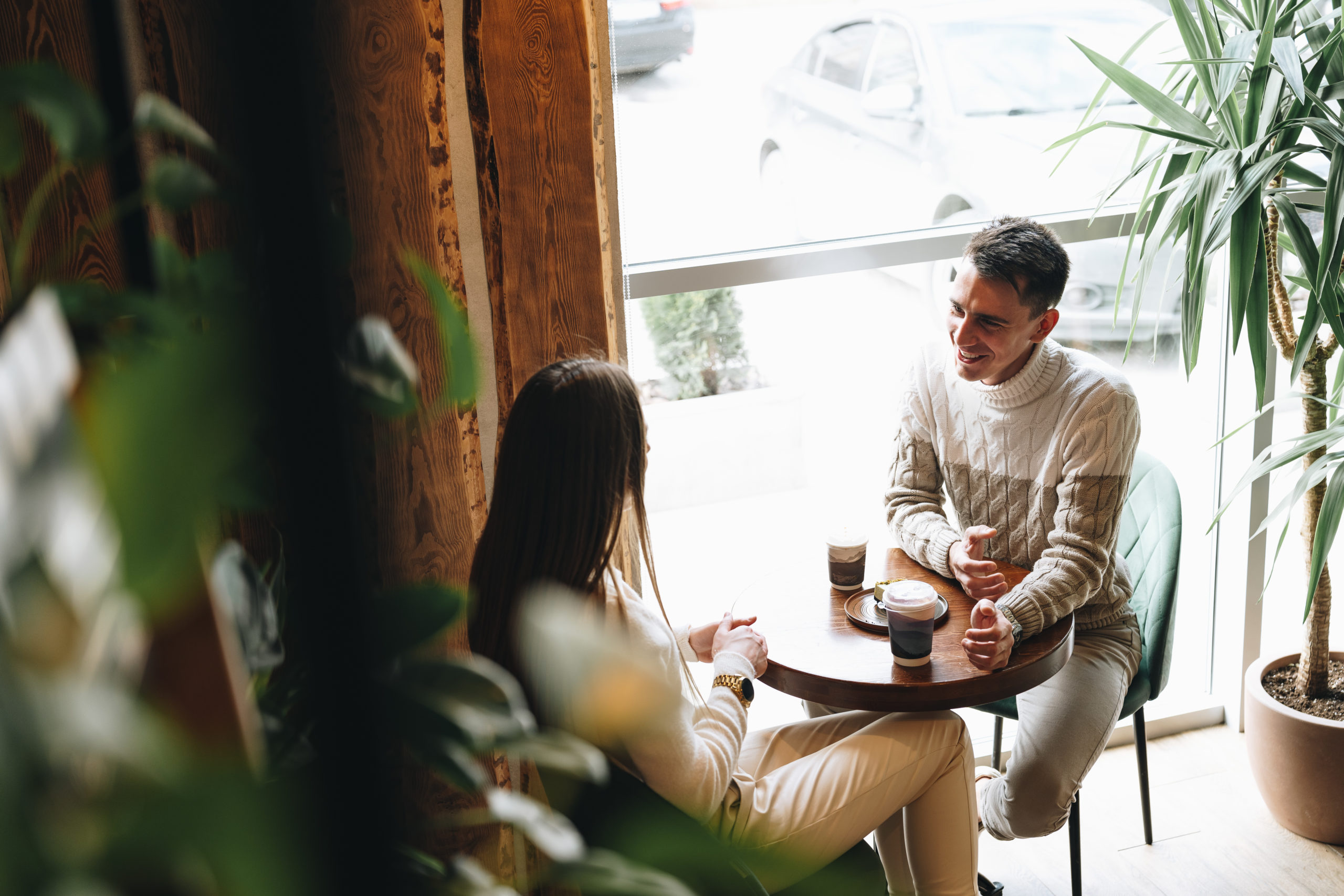 A man and woman are seated at a table engaging in conversation. They appear to be in deep discussion, leaning towards each other. The table is bare except for a few cups and plates. The man gestures with his hands while the woman listens intently.