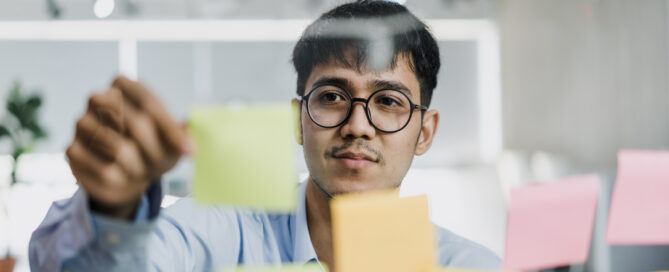 Focused man placing sticky notes on a glass board, symbolizing task management, organisation challenges, or cognitive planning in a work setting.