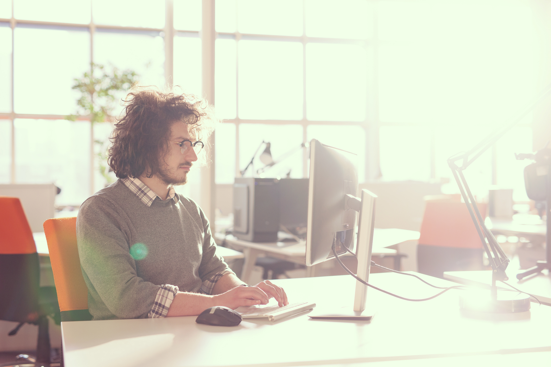 businessman working using a computer in startup office A man with curly hair and glasses sits at a desk working on a computer in a bright office with large windows.