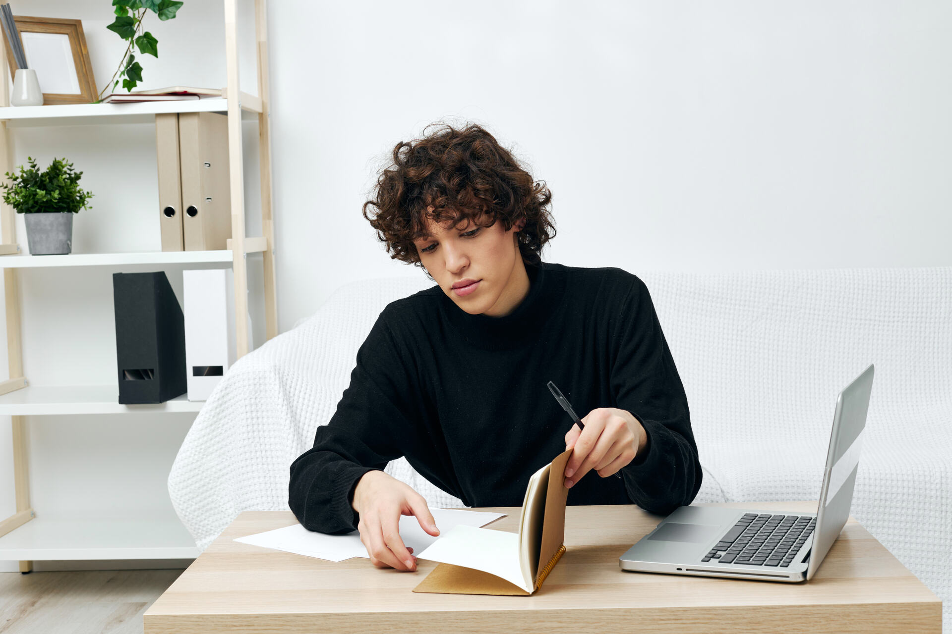 man sitting on the couch at the table in front of a laptop communication Person with curly brown hair in black sweater writing in notebook at wooden desk with laptop and white shelving unit with plants in background.