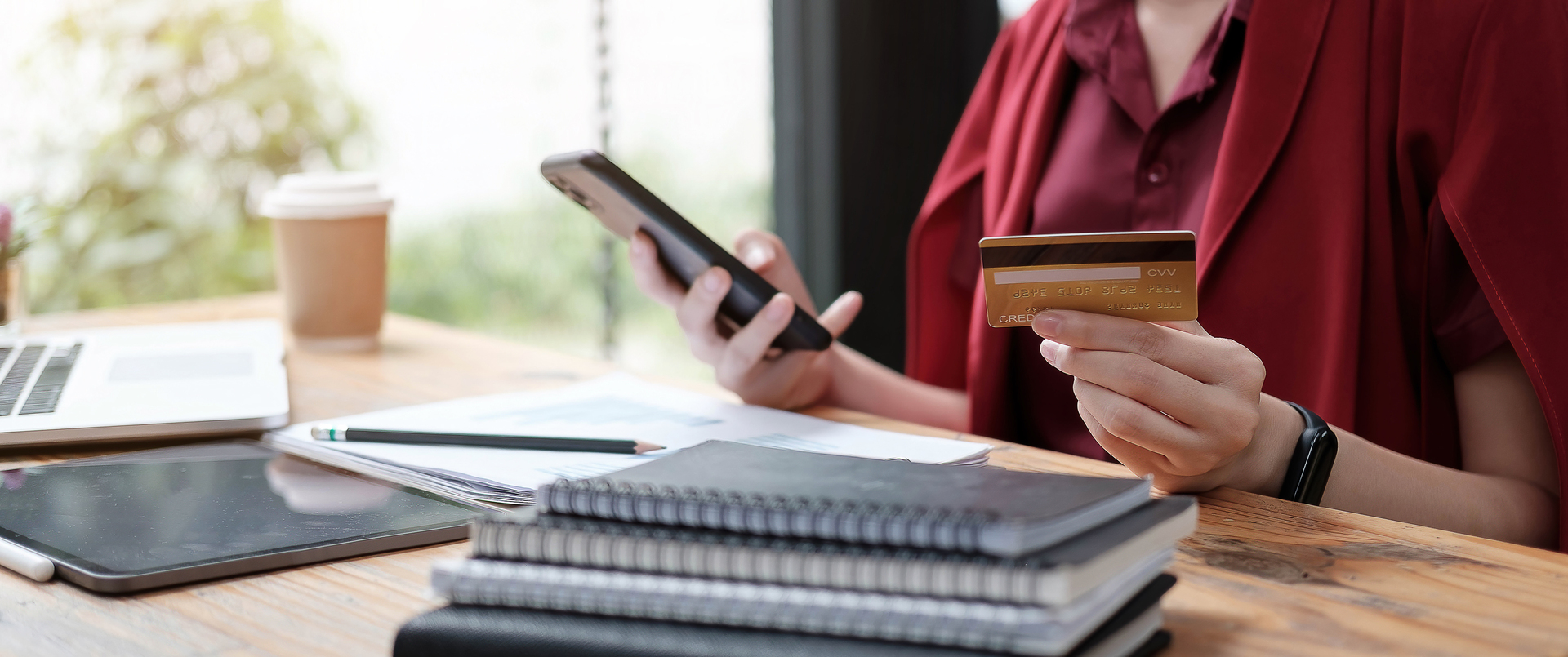 Online payment woman’s hands holding smartphone and using credit Person in red shirt holding smartphone and credit card at desk with laptop, notebooks, and coffee cup, making an online purchase.