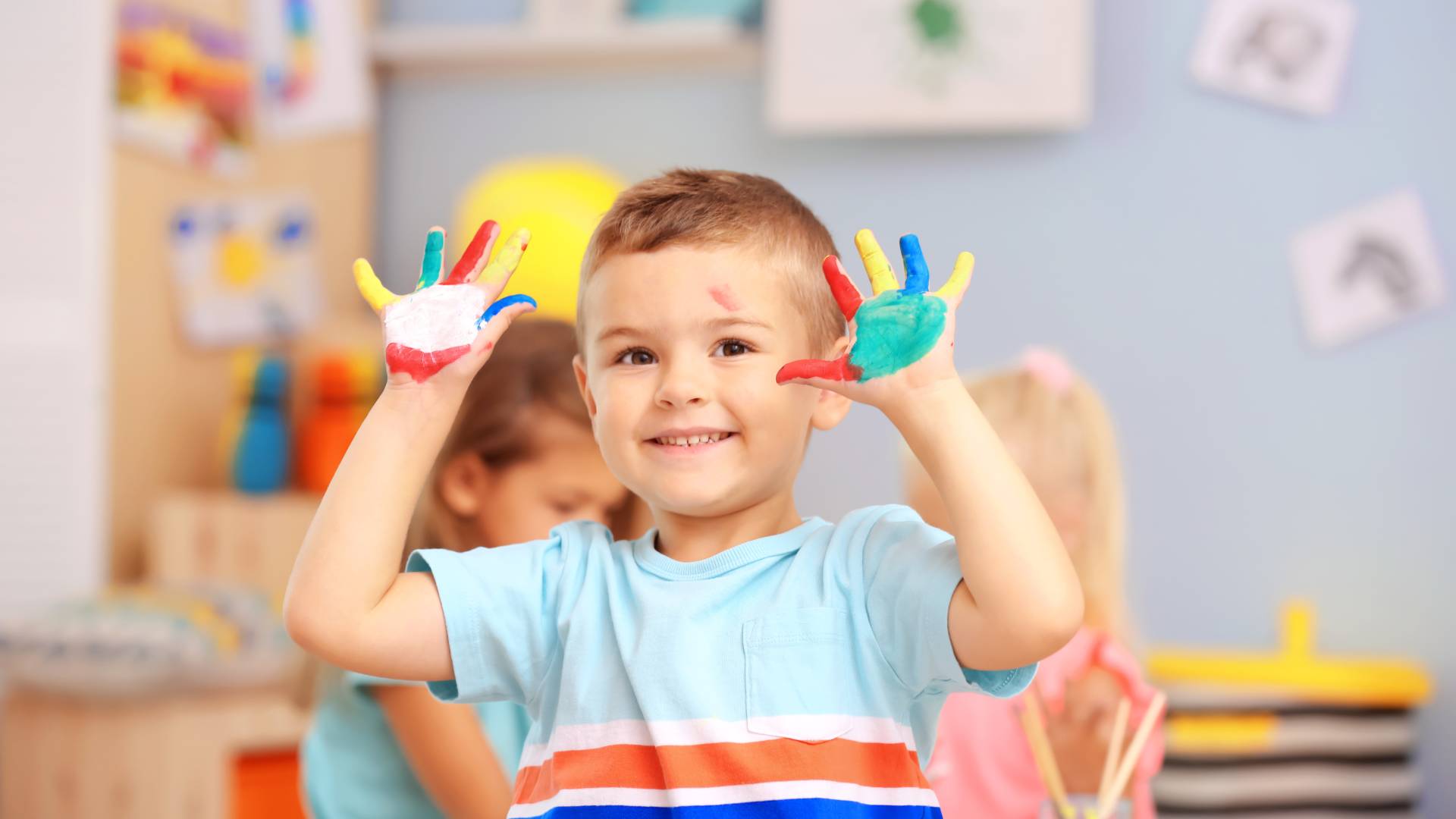 AdobeStock_216240694 a child in a classroom with multiple colours of paint on his hands, smiling