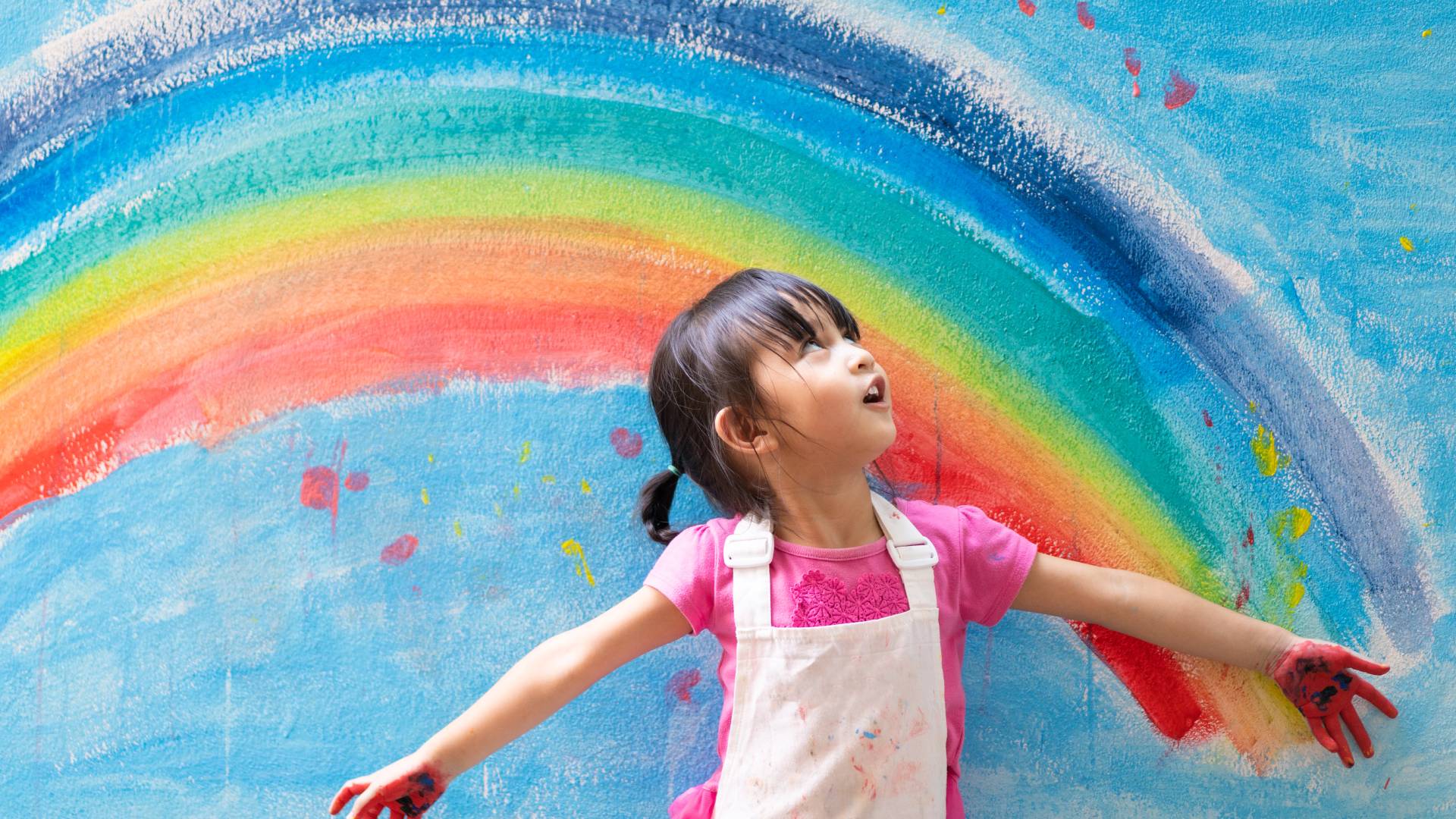 AdobeStock_369279046 a child with paint on her hands looking up into the sky against a backdrop of a painted rainbow on the wall behind her
