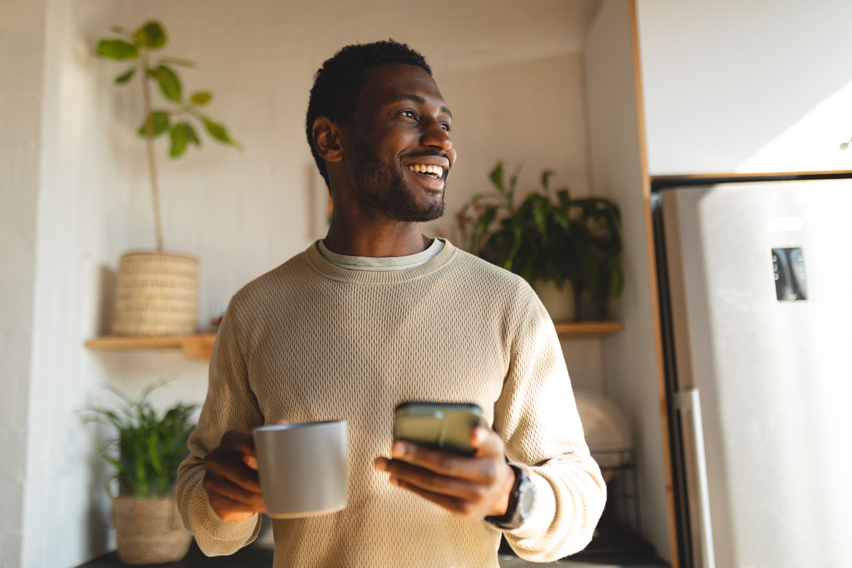 Image 2 a man in a cream jumper standing and looking into the distance smiling while holding his phone and a mug