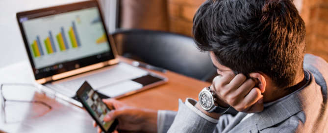 A man procrastinating on his phone sitting at his desk with an open laptop